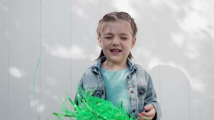 Cute happy girl dark hair smiling and celebrating saint patrick's day waving with green pom poms wearing a denim jacket white wall background