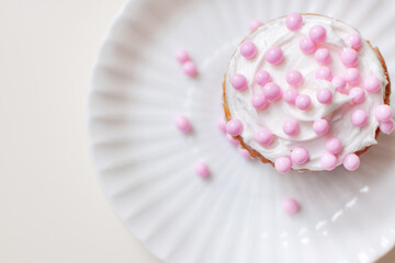 Vanilla cupcake with white cream frosting and round pink sprinkles, on white plate and surface, overhead view