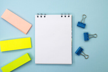 On a blue background, among paper clips and colored stickers, there is a notebook with a white blank sheet. Business and Education concept