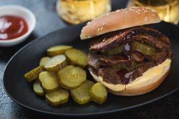 Black plate with brisket sandwich, pickles and beer mugs in the background, close-up, selective focus