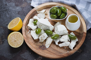 Round wooden serving tray with feta cheese, green olives and olive oil, studio shot on a brown stone background