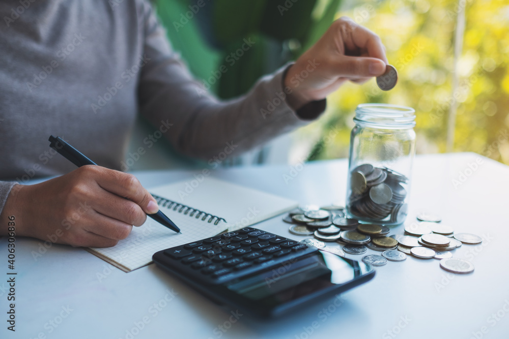 Wall mural Closeup image of a woman putting coins in a glass jar , calculating and taking note for saving money and financial concept