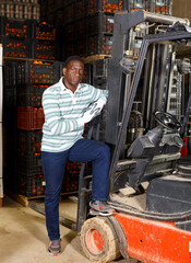 Fototapeta na wymiar Portrait of confident adult African American man standing near forklift at fruit warehouse