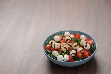 Blue bowl of salad with mozzarella, cherry tomatoes and kale, seasoned with balsamic sauce on walnut wood table