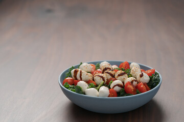 Blue bowl of salad with mozzarella, cherry tomatoes and kale, seasoned with balsamic sauce on walnut wood table
