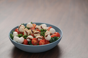 Blue bowl of salad with mozzarella, cherry tomatoes and kale, seasoned with balsamic sauce on walnut wood table