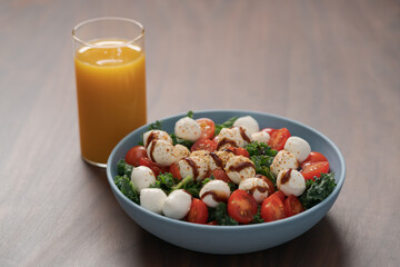 Blue bowl of salad with mozzarella, cherry tomatoes and kale, seasoned with balsamic sauce on walnut wood table with glass of orange juice