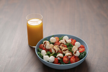 Blue bowl of salad with mozzarella, cherry tomatoes and kale, seasoned with balsamic sauce on walnut wood table with glass of orange juice