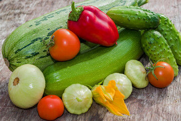 A pile of fresh vegetables on a wooden table. Harvest green zucchini, cucumbers, squash, red tomatoes, sweet peppers, illuminated by the sun.