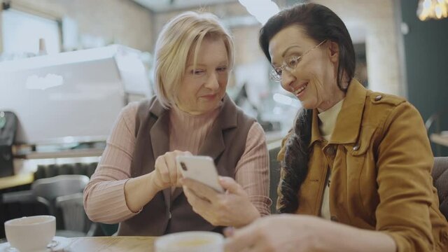 Modern elderly friends watching photos on smartphone, sitting in cafe together