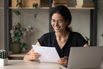Smiling millennial female in spectacles read paper documents work on computer online at home office. Happy young Caucasian woman in glasses consider paperwork use laptop for distant job or studying.