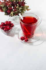 Autumn red herbal tea in transparent mug with hawthorn berries, rose hip, dry leaves in sunlight on white wood board, copy space, vertical, closeup.