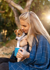 Nice laughing blonde girl in holding little dog jack russel and cup in hands. happily spending time on picnic in park covered blue blanket on sunset autumn forest background