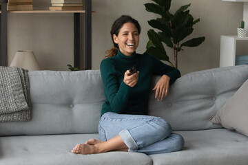 Overjoyed millennial Caucasian woman sit relax on sofa in living room enjoy cozy domestic weekend. Happy young 20s female rest on couch at home laugh watching TV program. Entertainment concept.