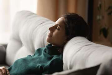 Calm young woman sit rest on cozy sofa at home sleep or take nap doing lazy weekend indoors....