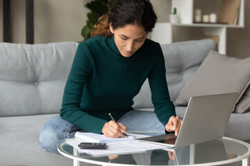 Focused young Caucasian woman manage family budget write expenses pay bills taxes online on laptop. Concentrated female take care of expenditures, make payment on computer. Saving concept.