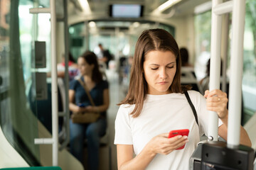 Fototapeta premium Young woman browsing and typing messages on phone on way to work in modern streetcar ..