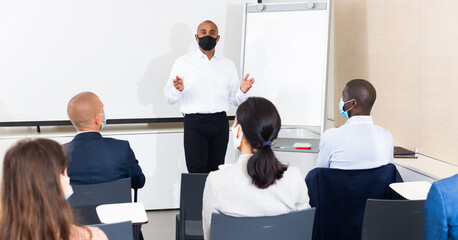 Confident Hispanic business coach wearing protective mask lecturing to group of businesspeople....