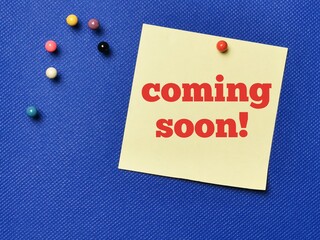 Text COMING SOON written on yellow paper note with pin board over blue background.Business concept.