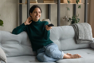 Happy young Caucasian woman sit relax on sofa watch TV switch channels on remote controller....