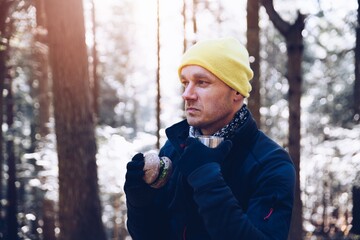 Handsome man tourist in yellow cap sitting on tree stump, eating hamburger and resting on the trail.