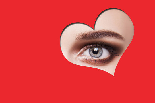 Young beautiful woman with smoky eyes makeup looking at camera through red heart shape. indoor studio shot isolated on red background.