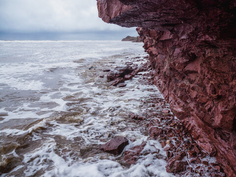 A cliff above the sea with a narrow coastline. Waves with white foam roll on the rocky shore. Tersky coast