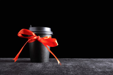 Black paper disposable takeout drink cup with bow made of gift red ribbon on dark surface and black...