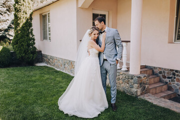 The groom in a gray checkered suit and the blonde bride in a white lace dress with a long veil are...
