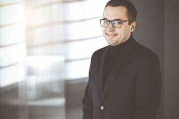 Portrait of a friendly middle aged businessman in a dark blue suit, standing straight in a sunny office. Concept of success in ba usiness nowadays