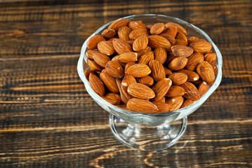 Almonds in a transparent glass bowl on an old shabby board. Nuts on a brown wooden table.