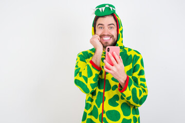 Joyous Young caucasian man wearing a pajama standing aga poses with mobile phone device, types text message on modern smartphone, watches funny video during free time, enjoys good internet connection,