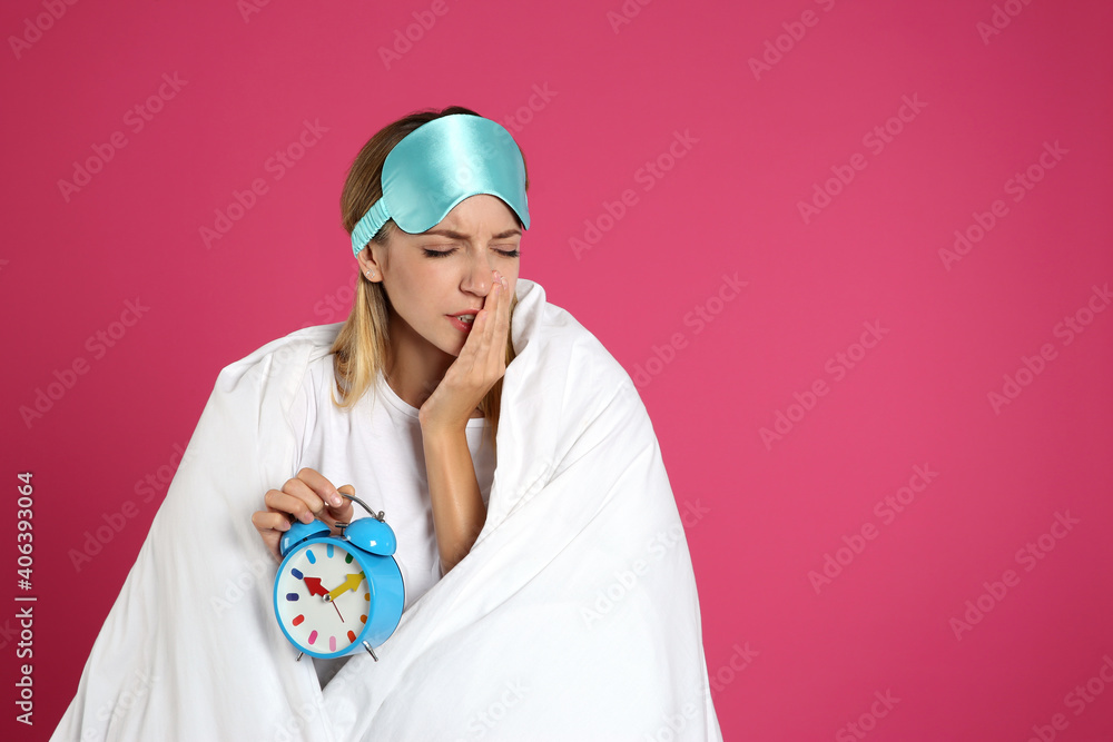 Wall mural Young woman in sleeping mask wrapped with blanket holding alarm clock on pink background. Space for text