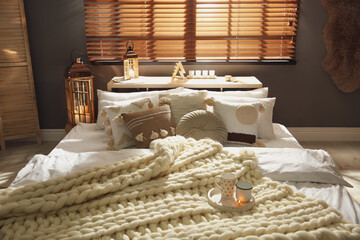 Bed with cozy knitted blanket and cushions near window in room. Interior design