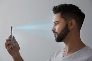 Young man unlocking smartphone with facial scanner on grey background. Biometric verification