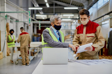 Mid adult engineer and male worker wearing face masks while examining paperwork and woodworking factory.