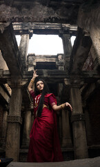 Portrait of beautiful Indian girl in heritage stepwell wearing traditional Indian red saree, gold...