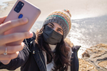 Hermosa chica con mascarilla y gorro haciendo selfie en invierno junto a la playa