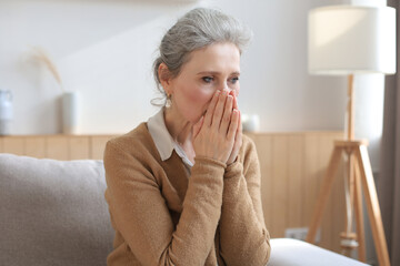 Stressed middle aged woman sit on sofa in living room, lost in thought.