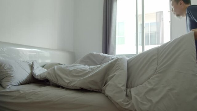 Asian man making the bed in his condo in the morning with sunshine coming in the window - slow motion