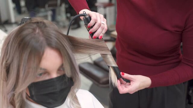 the hands of the hairdresser pinch a strand of hair with a straightener, stretch and scroll to curl a lock on the girl's long blond hair in front of a mirror in a protective mask