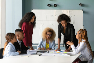 Obraz premium Focused multiethnic diverse colleagues coworkers gather at desk in office boardroom discuss company paperwork together. Multiracial businesspeople talk brainstorm at team meeting at workplace.