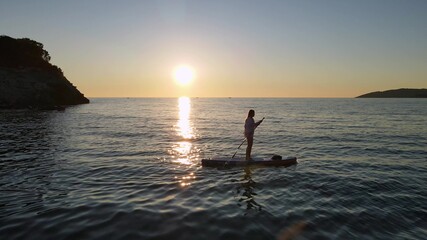 Silhouette of woman rowing SUP-board with paddle along sea with rippling waves against clear sky at sunset reflecting on water