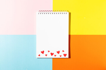 White notepad with red cardboard hearts on pink yellow blue and orange background copy space