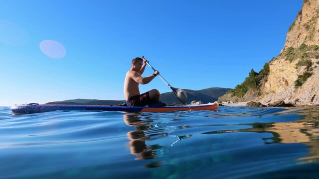 Young shirtless man rows contemporary SUP-board with paddle along azure sea water to old cliffs on sunny day low angle shot
