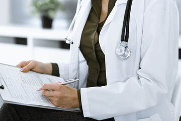 Unknown woman-doctor writing something at clipboard while sitting at the chair, close-up. Therapist wearing green blouse at work is filling up medication history record. Medicine concept