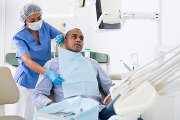 Obraz premium Portrait of adult hispanic man sitting on dental chair while woman dentist preparing him for medical examination in clinic