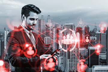 Obraz premium Handsome Caucasian HR director at international company is looking for highly qualified specialists to recruit by using smartphone. Social media hologram icons over Kuala Lumpur.