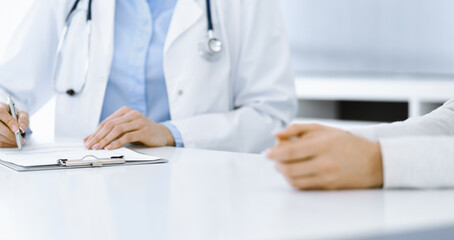 Unknown woman-doctor and female patient sitting and talking at medical examination in clinic, close-up. Therapist wearing blue blouse is filling up medication history record. Medicine concept