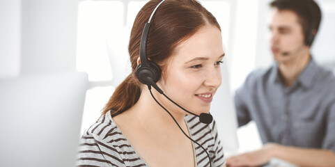 Naklejka premium Casual dressed young woman using headset and computer while talking with customers online. Group of operators at work. Call center, business concept